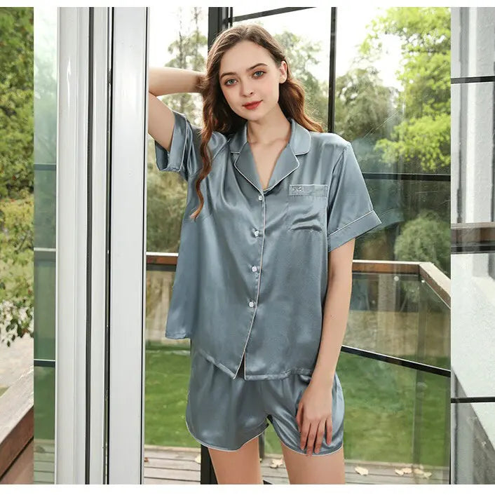 Woman wearing a gray satin pajama set standing by a glass door with a view of greenery outside.