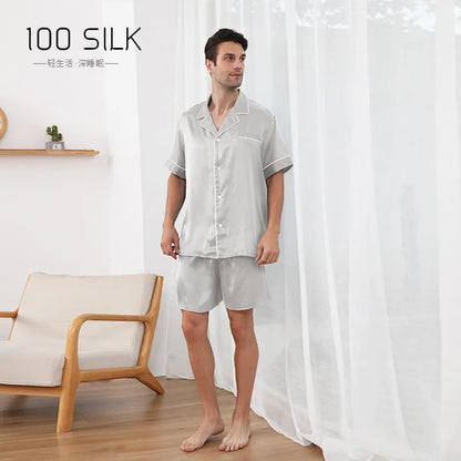 Man wearing a light gray silk pajama set standing in a room with a white curtain and wooden chair.