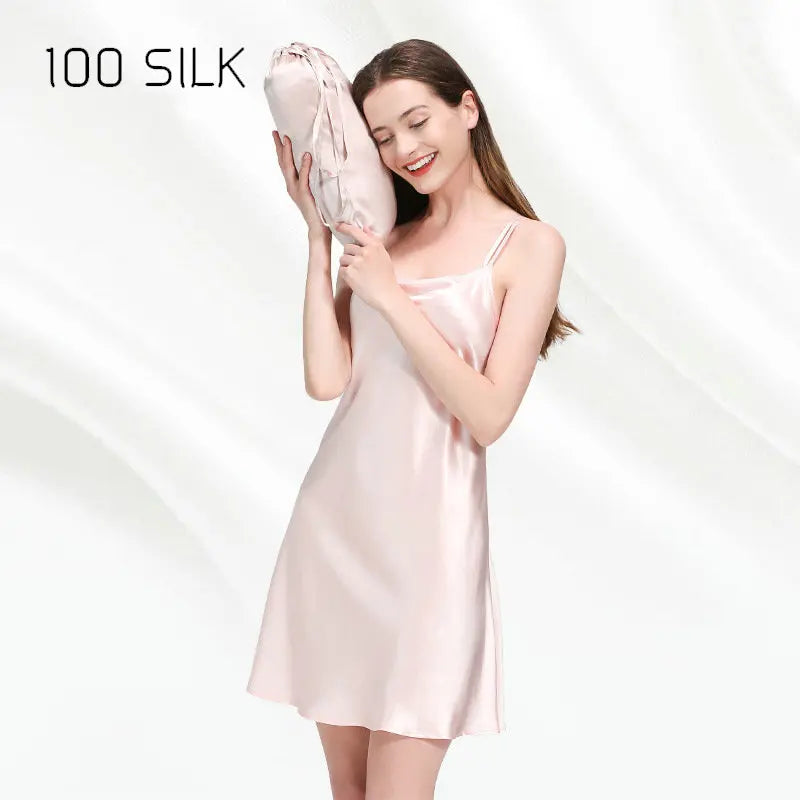 Woman wearing a pink satin slip holding a matching pillow with '100 SILK' branding.