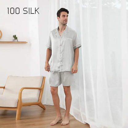 Man wearing a light gray silk pajama set standing in a room with a wooden chair and white curtains.