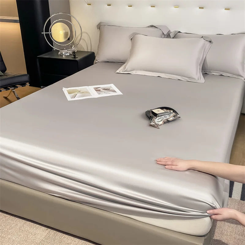 Person adjusting a silver satin sheet on a bed with a nightstand and lamp in the background.
